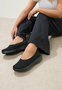 Schwarze Slipper mit einem strukturierten Netzoberteil und einer gepolsterten schwarzen Sohle. Das Design ist schlank und niedrigprofilig, geeignet für die lässige Freizeit.
