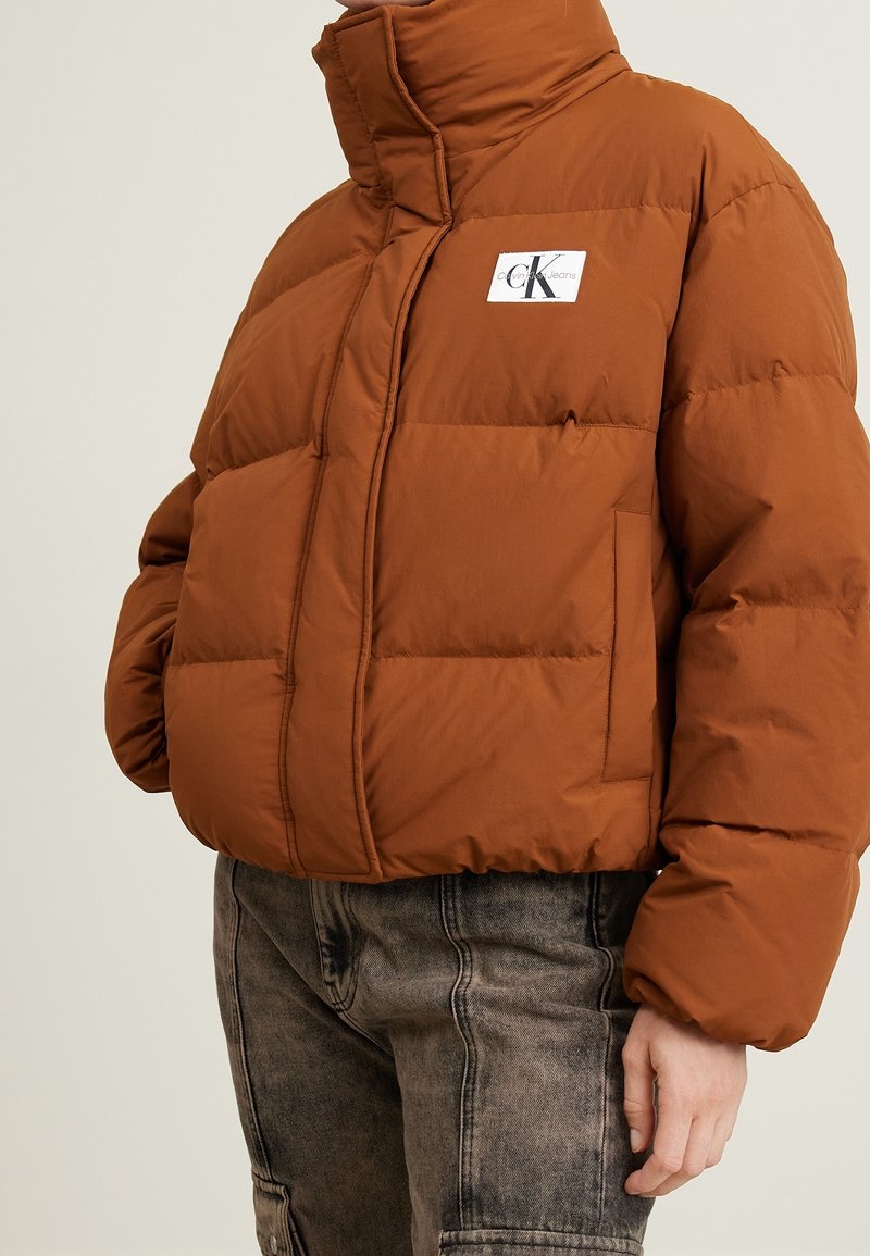 Doudoune marron avec col montant et logo Calvin Klein Jeans sur la poitrine, portée avec un jean noir délavé.