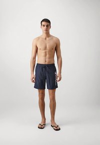 Temno modre plavalne kratke hlače z vrvico v pasu in detajlom logotipa na strani. Nošene s črnimi natikači. Model je brez majice in stoji na nevtralni podlagi.