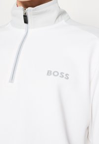 Pull-over blanc à demi-fermeture éclair avec col texturé et fermeture éclair grise, portant le logo "BOSS" sur la poitrine.