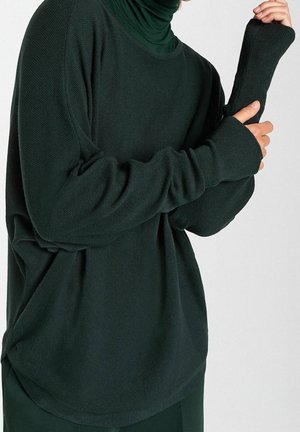 Pull en maille vert foncé avec une coupe décontractée, présentant des manches longues, un ourlet arrondi et des détails texturés sur le tissu.