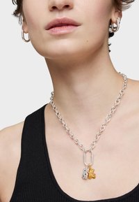 Srebrny naszyjnik łańcuszkowy z dużymi, wzajemnie tresującymi się ogniwami oraz dwoma zawieszkami: srebrnym misiem i złotym liściem. Tło jest neutralne.