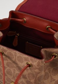 Borsa a secchiello in tela marrone con rifiniture in pelle e dettagli rossi. Dotata di accessori in oro, una tasca interna con cerniera e un interno foderato in bordeaux.