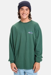 Groene langarmige shirt van katoen met een ronde hals en een logo-opdruk op de borst. G gedragen met lichtblauwe jeans en een marineblauwe beanie.