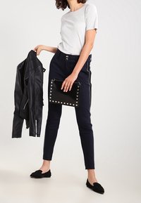 En kvinna med lockigt hår har på sig en kortärmad topp och marinblå byxor, håller en svart läderjacka i ena handen och en nitarprydd clutch i den andra.