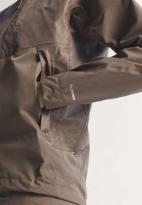 Veste marron imperméable avec poches latérales zippées et inscription "Climaproof" sur la manche, montrée avec la main dans la poche.