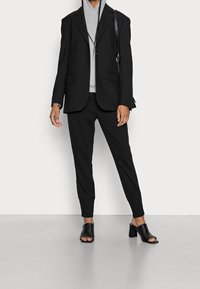 Person in einem schwarzen Blazer, schwarzen schmal geschnittenen Hosen und schwarzen offenen High-Heels, die eine schwarze Schultertasche trägt und geflochtenes Haar hat.