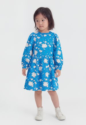 Mladé dievča s čiernymi vlasmi, oblečené v modré šaty s bielymi vzormi oblakov a hviezd a bielymi vysokými teniskami, stojace na bielom pozadí.