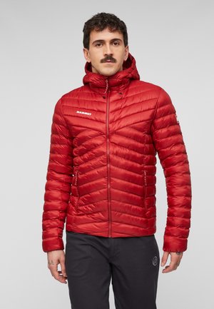 Mann mit Schnurrbart, der eine rote gesteppte Kapuzenjacke und schwarze Hose trägt, steht vor einem schlichten hellen Hintergrund.
