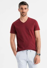 T-shirt a v in color bordeaux in cotone, maniche corte, indossato con pantaloni azzurri chiari. Presenta una vestibilità rilassata e dettagli di cucitura minimi.