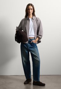 Mujer de pie contra una pared blanca, vistiendo una camisa de cuadros sobre una camiseta blanca, jeans azules, zapatos oscuros, sosteniendo un bolso de ante marrón oscuro con un brazo.