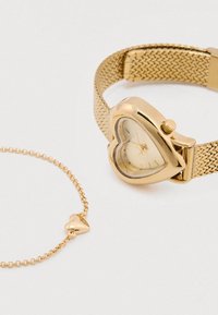 Kullavärvi südamekujuline kell võrgust rihmaga; läbipaistev sihverplaat minimaalse arvuga märgistega. Täienduseks sobib samas stiilis südamekujuline ripatsiga käevõru.