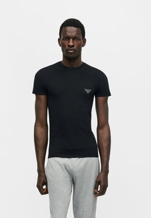 Sort tætsiddende T-shirt med korte ærmer og lille logo på brystet båret af stående mand med grå joggingbukser mod ensfarvet baggrund.