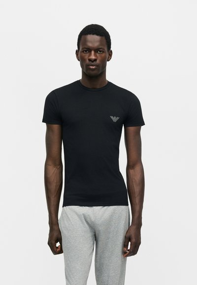 Μαύρο στενό T-shirt με κοντά μανίκια και μικρό λογότυπο στο στήθος, φορεμένο από όρθιο άνδρα με γκρι φόρμα, σε απλό φόντο.