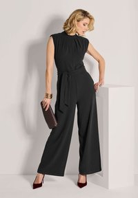 Vrouw in een mouwloze zwarte jumpsuit met een taillekoord, die een geweven clutch vasthoudt, gouden armbanden en bordeauxrode hakken draagt, leunend op een witte zuil.