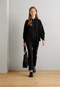 Černá pruhovaná košile s knoflíky, černé slim-fit kalhoty, černé boty a černá taška přes rameno, kráčející po světlé dřevěné podlaze proti šedé zdi.