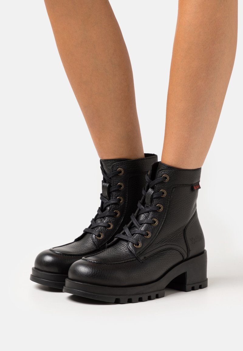 Bottines en cuir noir avec lacets, talons épais et semelles robustes portées sur des jambes nues sur un fond blanc uni.