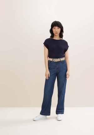 Top corto a righe blu scuro, jeans in denim blu a gamba larga a vita alta, cintura beige con fibbia, sneakers bianche. Sfondo neutro.