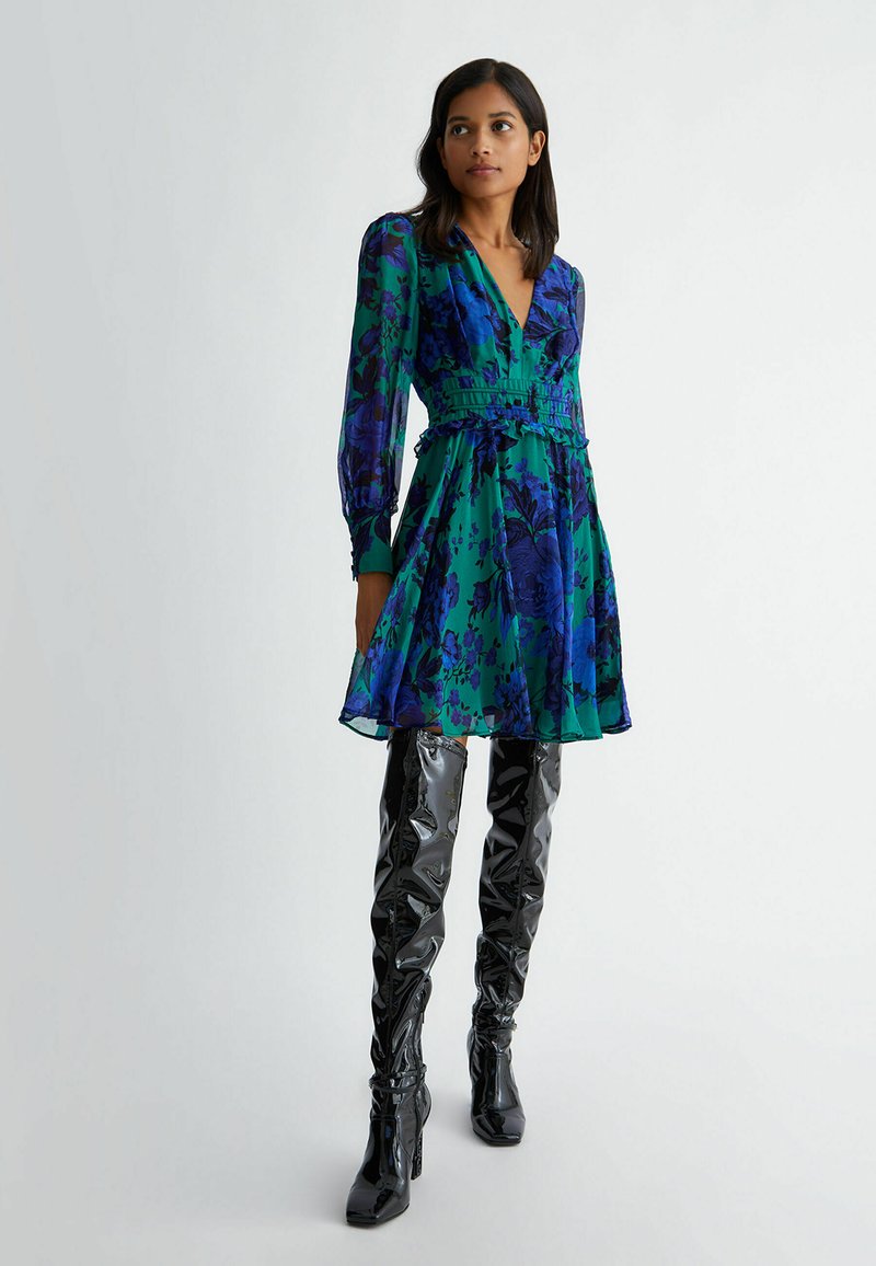 Abito floreale in verde e blu con scollatura a V, maniche lunghe, vita arricciata e orlo svasato, abbinato a stivaletti neri lucidi fino alla coscia.