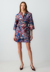 Krótka sukienka kimono z kolorowego jedwabiu, ozdobiona wzorem fal, z paskiem w talii i szerokimi rękawami. Paleta kolorów obejmuje róż, niebieski i zielony.