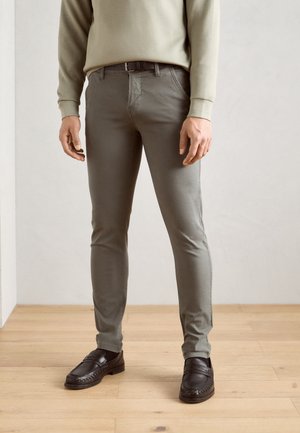 Mann trägt schmale graue Hose, schwarzen Gürtel, beige Langarmshirt und schwarze Leder-Loafer, steht auf Holzboden.