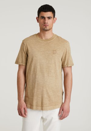 Camiseta de manga corta beige hecha de tejido texturizado, con cuello redondo y un pequeño parche de logo en el pecho.