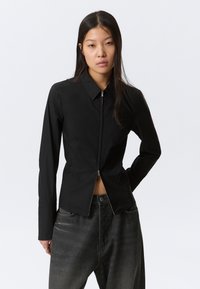 Femme portant un haut noir ajusté à manches longues avec fermeture éclair et un jean gris foncé ample, debout avec une main derrière le dos.