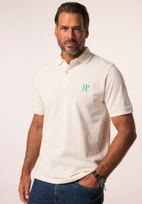 Kremowa koszulka polo z dwiema guzikami, krótkimi rękawami i turkusowym logo "JP 1880" na lewej piersi. Noszona z niebieskimi dżinsami i czarnym zegarkiem.