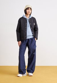 Schwarze gepolsterte Jacke, hellblaues Hoodie, marineblaue Cargo-Hosen und weiße Sneaker. Enthält eine gestrickte beige Mütze und sichtbare Logos auf den Hosen.