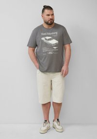 Grijze katoenen T-shirt met een Ford Mustang-graphic, gecombineerd met lichtcrème shorts en beige sneakers met donkere accenten en veters.