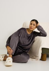 Ensemble de loungewear en velours gris avec des motifs étoilés, comprenant des manches longues et un pantalon. Pantoufles blanches en fausse fourrure avec des accents dorés en forme de cœur.