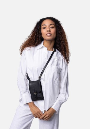 Schwarze Leder-Umhängetasche mit einem Frontklappe, abnehmbarem Schulterriemen und Schnallendetails. Das Modell trägt ein weißes Hemd und Hose vor einem neutralen Hintergrund.