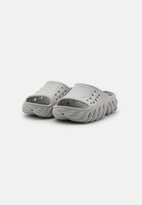 Grå slip-on klädda träskor gjorda av texturerad gummi med utskurna ventilationshål och en vågig sula design för förbättrat grepp och komfort.