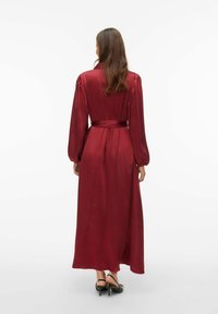 Długa bordowa sukienka z paskiem w talii, bufkami na rękawach i gładką, satynową teksturą, o płynącej sylwetce i długości do kostek.