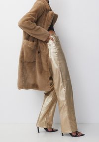 Manteau en fausse fourrure marron avec de grandes poches, associ é à un pantalon large brillant doré. Des talons noirs à brides complètent le look.