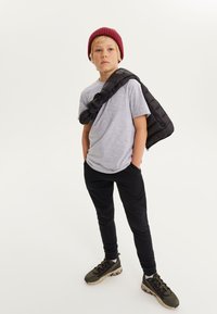 T-shirt gris à manches courtes, veste matelassée noire portée sur l'épaule, pantalon noir, baskets vert olive avec des semelles texturées, bonnet en maille rouge.