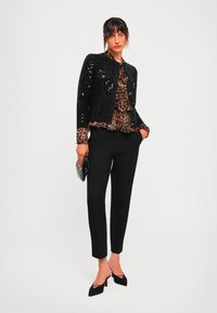 Mujer vistiendo una chaqueta negra con lentejuelas, blusa de estampado de leopardo, pantalones negros ajustados, tacones negros, sosteniendo un pequeño clutch negro contra un fondo blanco.