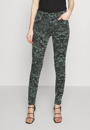 Kobieta w czarno-zielonych obcisłych spodniach z kieszeniami, czarnej bluzce i czarnych wysokich sandałach na cienkich paskach stojąca.