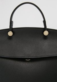 Sac à main en cuir texturé noir avec une poignée supérieure et des clous ronds dorés gravés du nom de la marque de chaque côté de la fixation de la poignée.