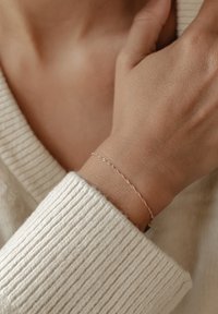 Pulsera delicada de oro rosa con un diseño retorcido, llevada en una muñeca por encima de un suéter de color crema con canalé. Presenta pequeños detalles texturizados.