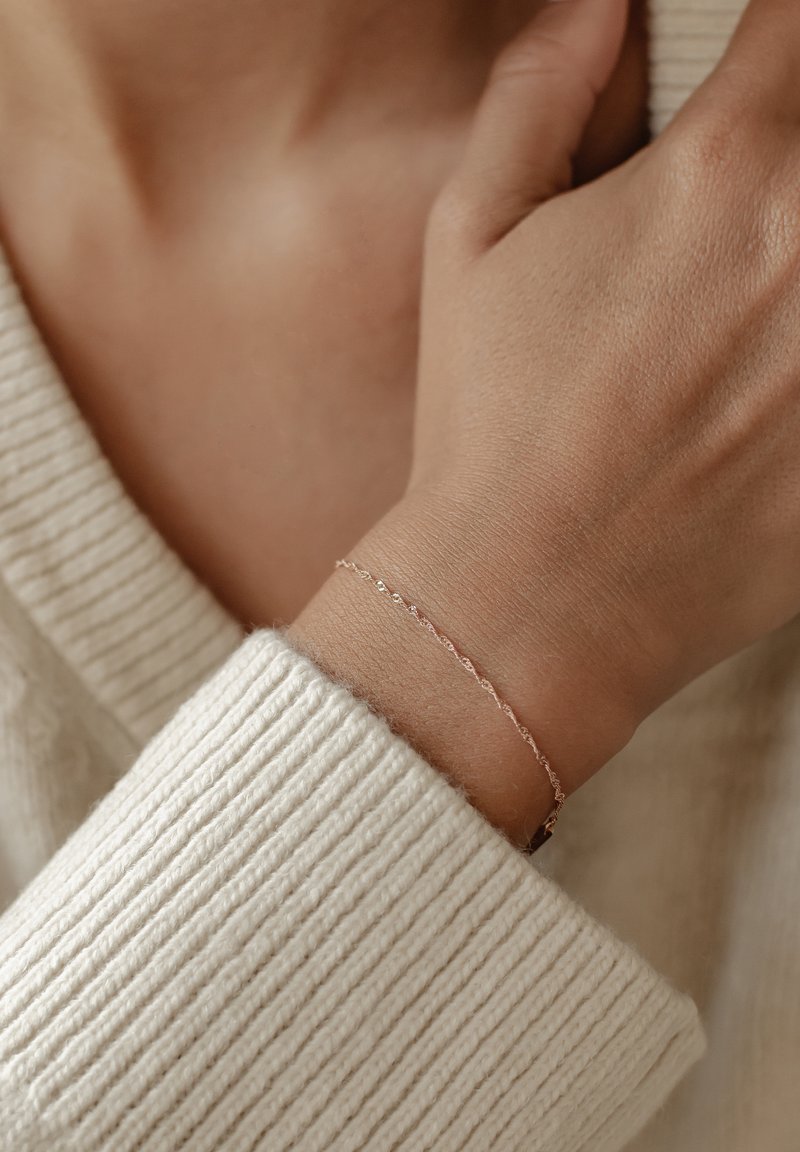 Pulsera delicada de oro rosa con un diseño retorcido, llevada en una muñeca por encima de un suéter de color crema con canalé. Presenta pequeños detalles texturizados.