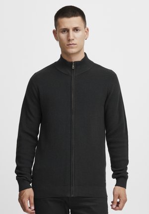 Cárdigan negro con cremallera, textura acanalada, cuello alto y mangas largas. Presenta puños ajustados y un acento de logo en el pecho.