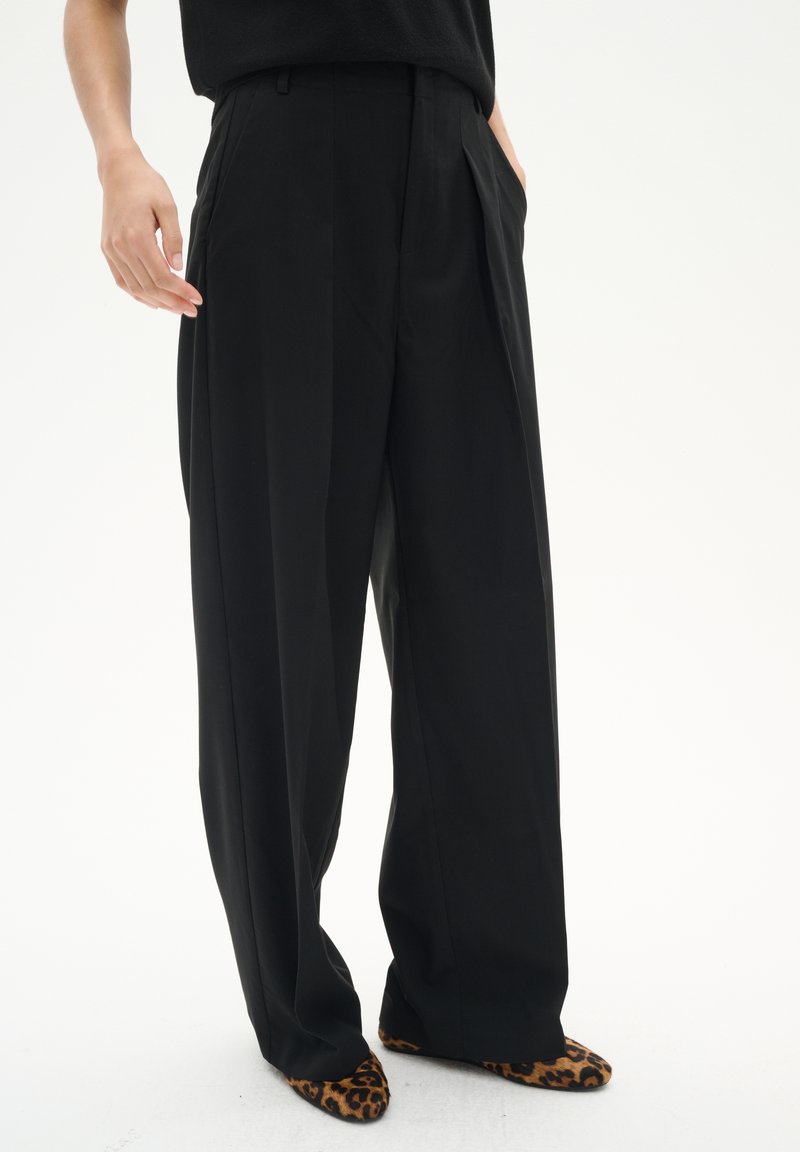 Personne portant un pantalon noir ample associé à des chaussures pointues avec imprimé léopard, debout devant un fond blanc uni.