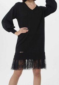 Svart stickad klänning med v-halsringning, ballongärmar och en genomskinlig spetskant. Har varumärkes detaljer i midjan. Texturerad och lager-på-lager design.