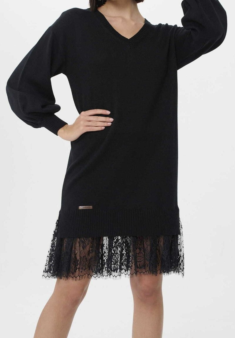 Svart stickad klänning med v-halsringning, ballongärmar och en genomskinlig spetskant. Har varumärkes detaljer i midjan. Texturerad och lager-på-lager design.