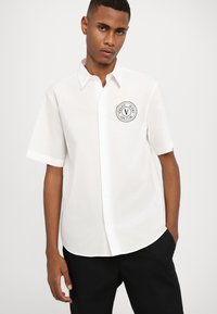 Chemise blanche à manches courtes en tissu léger, dotée d'un logo noir circulaire sur la poitrine gauche, d'un col classique et d'une fermeture à boutons sur le devant.
