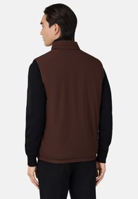 Hnedá bezrukávová vesta s vysokým golierom; jemná, hladká textúra; čierna košeľa s dlhým rukávom pod ňou; elastický lem; jemné švy.