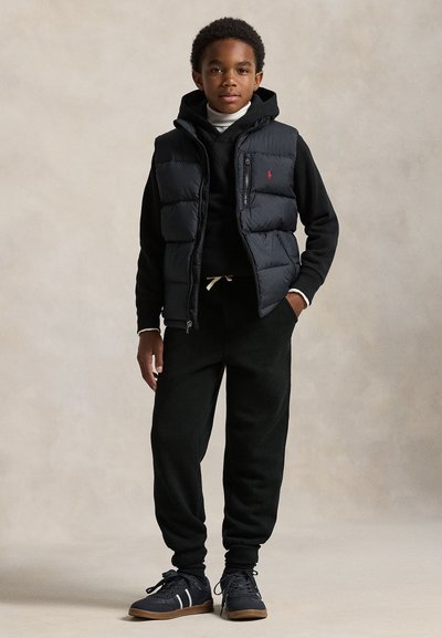 Chlapec v černé péřové vestě přes černou mikinu s kapucí a tepláky, s černými teniskami, na neutrálním vnitřním pozadí.