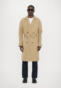 Trench coat bej cu două rânduri de nasturi, cu brâu, buzunare laterale și guler tăiat, asortat cu blugi închis la culoare și pantofi negri.