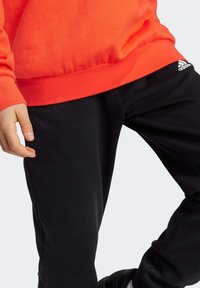 Sudadera naranja de mangas largas con dobladillo acanalado, combinada con pantalones de chándal negros con el logo de Adidas, diseño elegante y material suave.